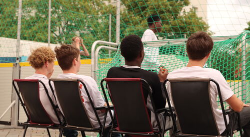 Vier Jugendliche sitzen auf Klappstühlen und sehen dem Fußballspiel zu.