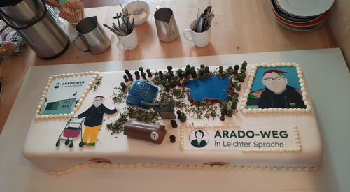 Eine rechteckige Torte mit weißem Zuckerguss ist verziert mit Figuren, Pyramide, Schokoriegel und Flächen mit schwarzer Schrift: Arado-Weg in Leichter Sprache