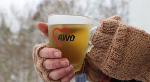 Teetasse mit AWO-Logo. Tasse wird mit zwei Händen in fingerlosen braunen Handschuhen gehalten.