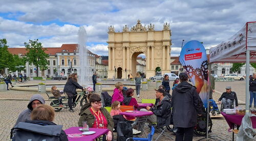 Luisenplatz Potsdam, das Brandenburger Tor im Hintergrund. Vorne Tische und Menschen die Kuchen essen und sich unterhalten. Ein Springbrunnen in der Mitte.