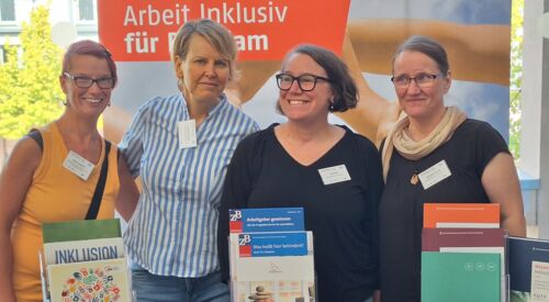 4 Frauen stehen nebeneinander am Stand des Netzwerk Arbeit inklusiv und lächeln in die Kamera. Vor ihnen auf dem Tisch Informationsmaterial zum Thema Arbeit mit Behinderung.