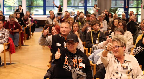 Großer Raum, Viele Teilnehmende sitzen auf Stühlen, lächeln und strecken den Daumen nach oben.