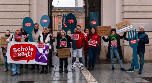 Mehrere Personen stehen vor dem Brandenburgischen Landtag. Sie halten Plakate hoch, auf denen steht: Schule satt.