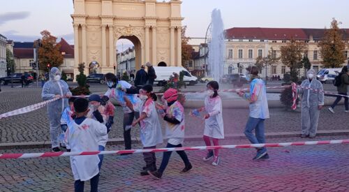 Der Luisenplatz mit Springbrunnen und brandenburger Tor im Hintergrund. Ein Absperrband wird von Menschen in Schutzanzügen / Masken gehalten sodass ein Rechteck umspannt ist, darin Mädchen in weißen Shirts bunt befleckt, auch der Steine / sind kunterbunt.